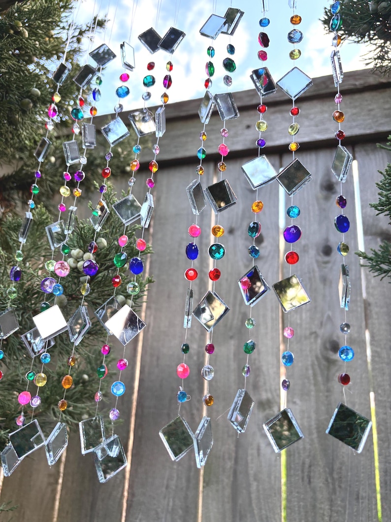 Puede incluir: Cadenas decorativas colgantes con cuadrados espejados y cuentas coloridas en tonos rosa, azul, verde, morado y naranja. Las cadenas cuelgan frente a una valla de madera y vegetaci&oacute;n.