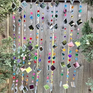 Puede incluir: M&oacute;vil decorativo colgante con hilos de cuentas de colores y piezas de vidrio espejado cuadradas. Las cuentas son de varios colores, incluyendo rojo, azul, verde y morado. El m&oacute;vil cuelga frente a una valla de madera.