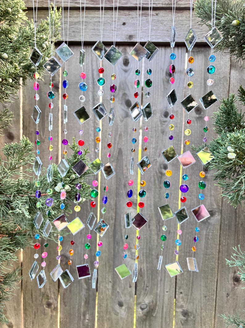 Puede incluir: M&oacute;vil decorativo colgante con hilos de cuentas de colores y piezas de vidrio espejado cuadradas. Las cuentas son de varios colores, incluyendo rojo, azul, verde y morado. El m&oacute;vil cuelga frente a una valla de madera.