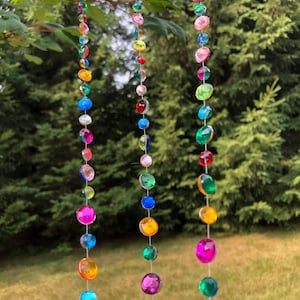 Puede incluir: Tres hilos de cuentas de colores y facetadas cuelgan de una rama de &aacute;rbol. Las cuentas son de varios tama&ntilde;os y colores, incluyendo rosa, azul, verde, amarillo y naranja. El fondo es una vista borrosa de &aacute;rboles verdes y hierba.