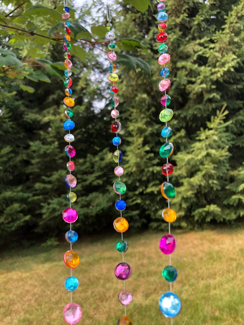 Puede incluir: Tres hilos de cuentas de colores y facetadas cuelgan de una rama de &aacute;rbol. Las cuentas son de varios tama&ntilde;os y colores, incluyendo rosa, azul, verde, amarillo y naranja. El fondo es una vista borrosa de &aacute;rboles verdes y hierba.