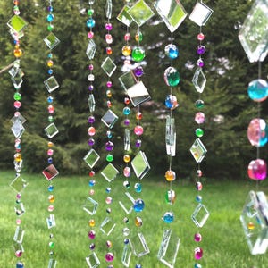 Puede incluir: Cadenas decorativas colgantes con piezas de vidrio transparente en forma de diamante y rectangulares, intercaladas con cuentas de colores en tonos rojos, naranjas, amarillos, verdes, azules y morados. Las cadenas cuelgan sobre un fondo verde borroso.