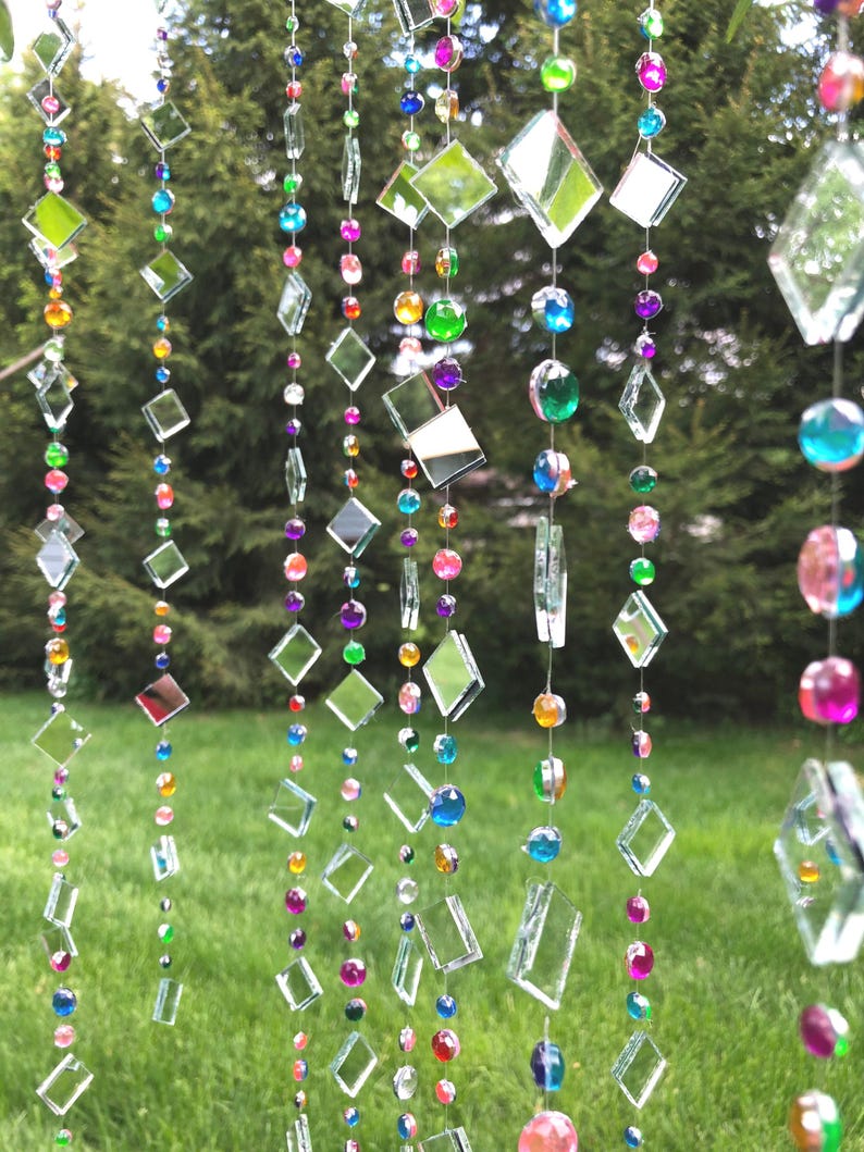 Puede incluir: Cadenas decorativas colgantes con piezas de vidrio transparente en forma de diamante y rectangulares, intercaladas con cuentas de colores en tonos rojos, naranjas, amarillos, verdes, azules y morados. Las cadenas cuelgan sobre un fondo verde borroso.