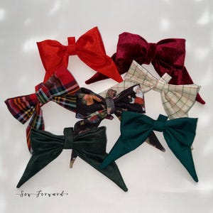 May include: A collection of seven fabric hair bows in various colours and patterns, including solid red, deep burgundy velvet, green velvet, a red and green plaid, a cream plaid, and a black bow with a Santa Claus print. Text reads Sew Forward.