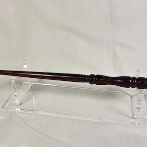 May include: A handcrafted, dark brown wooden wand with a polished finish. The wand features a detailed handle with carved rings and a tapered tip. It is displayed on a clear acrylic stand against a white background.