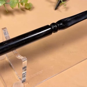 May include: A smooth, black, cylindrical wand featuring a tapered handle with a rounded pommel and a distinct decorative ring detail near the base. The wand is crafted with a polished finish and rests horizontally on a clear acrylic display stand.