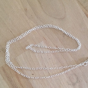 May include: A delicate silver chain necklace with a small clasp, lying on a light brown wooden surface. The chain is made of small, interconnected links. The necklace is a simple, elegant piece of jewelry.