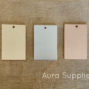 May include: Three rectangular paint chips with a hole punched in the top. The chips are in the colors cream, silver, and light pink. The text "Aura Supplies" is printed below the chips.