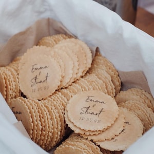 May include: A basket of round, pale brown biscuits with a decorative, scalloped edge. Some biscuits are printed with the names "Emma & Stuart" and the date "12.05.23". The basket is lined with white fabric and brown paper.