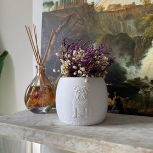 May include: A white ceramic plant pot featuring a detailed embossed bulldog design. The pot is filled with dried purple and white flowers. A glass diffuser bottle with reeds is in the background, along with a painting.