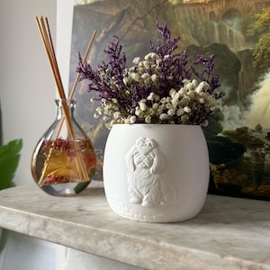 May include: A white ceramic vase with a dog relief, holding purple and white dried flowers. A glass diffuser with reeds and liquid is to the left. The vase is on a marble surface.