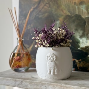 May include: A white ceramic vase with a dog relief, holding dried purple and white flowers. The vase is on a marble surface, with a glass diffuser bottle in the background. The vase is filled with dried flowers.