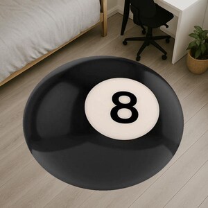 May include: A round black rug with a white circle featuring the number 8 in black. The rug is designed to resemble an eight ball from a game of pool. The rug is on a light wood floor.