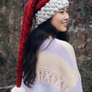 May include: A long, red and white Santa hat with a white pom-pom. The hat is made of a knitted material. The person is wearing a striped sweater with a light purple, cream, and light yellow color scheme.
