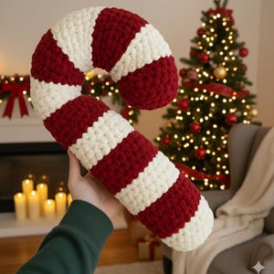 Puede incluir: Una almohada grande y hecha a mano con forma de bastón de caramelo con rayas rojas y blancas alternas. La almohada de felpa curvada se sostiene frente a un árbol de Navidad con luces y adornos. El bastón de caramelo es una decoración navideña festiva.