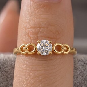 Könnte beinhalten: Ein goldener Ring mit einem zentralen, rund geschliffenen, klaren Edelstein in einer Vier-Krappen-Fassung. Das Band ist mit zwei symmetrischen, ineinandergreifenden, schleifenförmigen Metallgliedern auf beiden Seiten des Steins gestaltet.