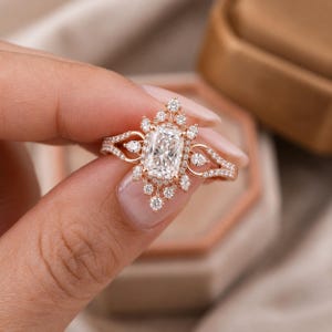 Könnte beinhalten: Ein Ring aus Roségold mit einem zentralen, im Radiant-Schliff geschliffenen klaren Stein, umgeben von einem Kranz aus kleineren runden und birnenförmigen Steinen. Die Ringschiene ist geteilt und mit Pavé-Diamanten besetzt.