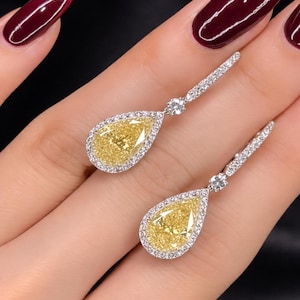 Può includere: Un paio di eleganti orecchini pendenti con gemme gialle a forma di pera, circondate da un alone di pietre più piccole e chiare. Gli orecchini sono incastonati in un metallo color argento.