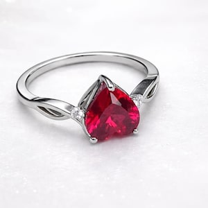 Könnte beinhalten: Ein silberner Ring mit einem herzförmigen, leuchtend roten Edelstein. Der Ring hat ein zartes, verdrehtes Banddesign mit kleinen, klaren Akzentsteinen auf beiden Seiten des herzförmigen Steins. Ein Schmuckstück.