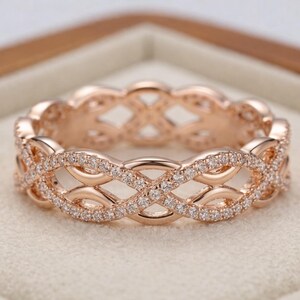 Puede incluir: Anillo de eternidad de oro rosa con un diseño entrelazado continuo. El anillo está incrustado con pequeñas piedras claras y brillantes. El anillo se muestra en una caja de joyería de tonos neutros.