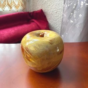 Pode incluir: Uma escultura decorativa de maçã com um acabamento brilhante e marmorizado, apresentando padrões em espiral em tons de amarelo dourado, castanho-claro e castanho-escuro. O objeto tem uma forma lisa e arredondada com um pequeno caule.