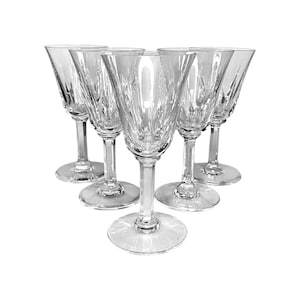 Op de afbeelding: Een set van vijf wijnglazen van helder glas, gerangschikt in een driehoekige formatie. Elk glas heeft een uitlopende kelk met verticale geslepen facetten aan de onderkant, een slanke steel en een ronde voet, vervaardigd uit transparant glas.