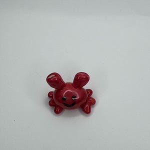 Puede incluir: Pequeña figura de cangrejo de cerámica roja brillante con una cara sonriente. El cangrejo tiene dos ojos grandes y redondeados y varias patas pequeñas. La figura es un objeto decorativo.