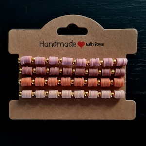 May include: A stack of handmade bracelets in shades of pink, peach, and brown, interspersed with gold beads. The bracelets are presented on a brown card, which has the text "Handmade with love". The overall aesthetic is earthy and neutral.