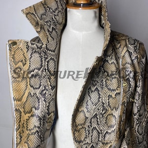 May include: A snakeskin-patterned jacket with a high collar and a zipper closure. The jacket is displayed on a mannequin. The color palette includes shades of brown, tan, and black, creating a realistic reptile skin effect. The jacket has gold-colored hardware.