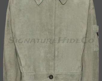 May include: A sage green suede jacket featuring a pointed collar, a button-front closure, and a small utility pocket on the left sleeve. The text SIGNATUREHIDECO is printed across the center of the jacket in a dark, semi-transparent font.