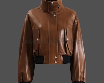 May include: A brown leather jacket featuring a high stand-up collar with a snap-button tab, a front zipper closure concealed by a placket with silver-tone snap buttons, two vertical flap pockets, and black ribbed knit cuffs and hem.