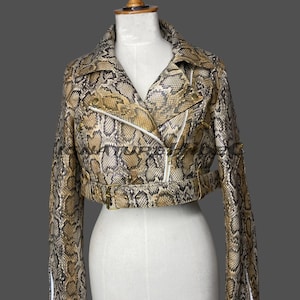 Könnte beinhalten: Eine kurz geschnittene Jacke mit einem Schlangenledermuster in Beige, Braun und Schwarz. Die Jacke verfügt über einen Reverskragen, einen asymmetrischen Frontreißverschluss, lange Ärmel mit Reißverschlussbündchen und einen Gürtel mit goldener Schnalle.