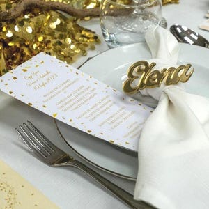 Può includere: Una tavola formale con un tovagliolo bianco tenuto da un anello portanome dorato "Elena". Un menu con testo dorato e coriandoli è su un piatto bianco. Una forchetta e un coltello sono sul tavolo. Il menu recita "10 luglio 2023".