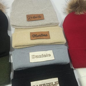 Puede incluir: Una pila de gorros personalizados en varios colores, incluyendo gris, beige, azul, negro y rojo. Cada gorro tiene un parche de cuero con un nombre: Roberto, Martina, Daniele y Gabriele. Algunos gorros tienen un pompón de piel sintética.