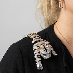 May include: A bejeweled tiger brooch with clear and green gemstones, and gold and silver accents. The brooch is draped over a black garment. The tiger's eyes are green, and the brooch is set with many small, clear gemstones.