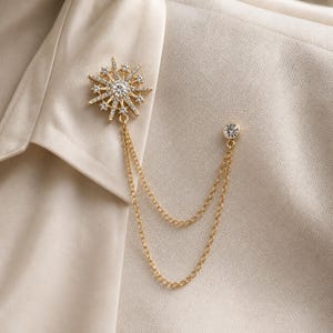 May include: Gold-toned brooch featuring a starburst design with clear crystals. The brooch has a central crystal and dangling chains. The pin is attached to a light-colored fabric, likely a blazer or jacket.