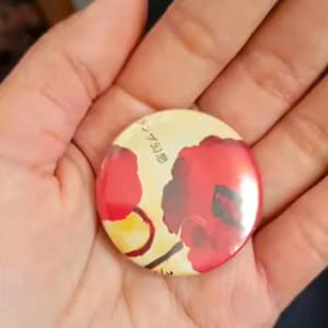 May include: A round button with a floral design featuring red flowers on a cream background. The button is held in a hand. Japanese text is visible on the button.