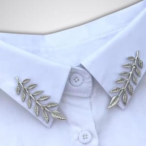 May include: Silver-colored leaf collar pins on a white dress shirt. The pins are shaped like branches with detailed leaves, adding a touch of nature-inspired elegance to the shirt's collar. The shirt has a classic button-down design.