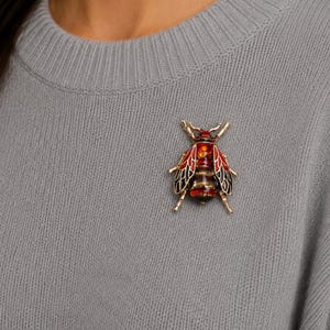 Peut inclure: Broche insecte dorée avec des détails en émail rouge, noir et ambre. La broche est épinglée sur un pull gris, mettant en valeur le design complexe et les couleurs vives du bijou.