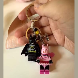 Puede incluir: Un llavero con dos figuras de Batman estilo Lego. Una es negra con un emblema amarillo, la otra es rosa con un emblema a juego y un tutú. El llavero tiene un anillo dorado y abalorios decorativos.