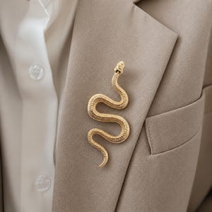 Könnte beinhalten: Eine goldfarbene Schlangenbrosche, die an einem beigen Blazer befestigt ist. Die Brosche weist detaillierte Schuppen und einen erhobenen Kopf auf. Der Blazer wird über einem weißen Hemd getragen, was einen Lagenlook erzeugt. Die Brosche verleiht dem Outfit einen Hauch von Eleganz.