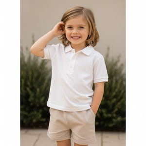 Pode incluir: Uma crian&ccedil;a vestindo uma camisa polo branca com gola e mangas curtas, combinada com shorts c&aacute;qui. A crian&ccedil;a est&aacute; sorrindo e com a m&atilde;o perto da cabe&ccedil;a. A roupa &eacute; simples e cl&aacute;ssica.