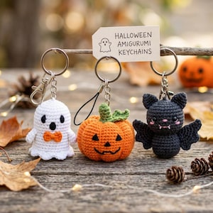 May include: Three crocheted amigurumi keychains featuring a white ghost with an orange bow tie, an orange pumpkin with a green stem, and a black bat with pink cheeks. A tag above reads HALLOWEEN AMIGURUMI KEYCHAINS with a ghost icon.