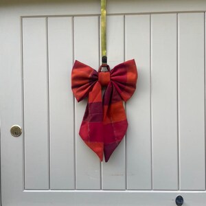 May include: A large fabric bow featuring a plaid pattern in shades of red, orange, and deep magenta. The bow is attached to a long, thin, metallic gold-colored vertical hook that extends upward, allowing it to be suspended from a door top.