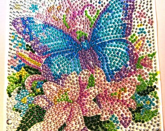 Puede incluir: Pintura de diamantes cuadrada que representa una mariposa con alas azules y moradas, rodeada de flores rosas y moradas y hojas verdes. La obra de arte está adornada con pequeños pedrería brillantes, creando un efecto brillante.