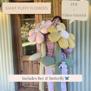 May include: A person holds giant, puffy fabric flowers with pink and yellow petals, a green stem, and a bee and butterfly. The text "GIANT PUFFY FLOWERS" and "Includes Bee & Butterfly" are displayed.