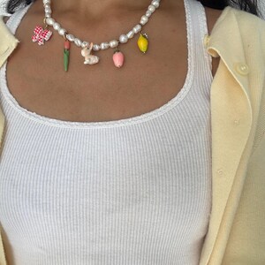 Puede incluir: Un collar de perlas con colgantes de colores. Los colgantes incluyen un lazo de cuadros rojos y blancos, un tulipán rosa, un conejito blanco, una fresa rosa y un limón amarillo.