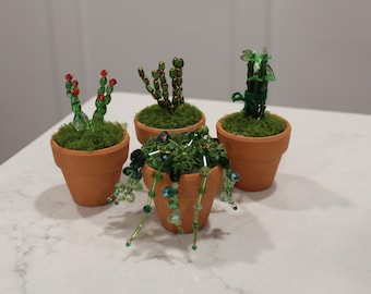 May include: Four miniature terracotta pots containing artificial plants crafted from green and red glass beads. Each pot is filled with green moss. The plants include a beaded cactus with red tips, a tall beaded succulent, a bamboo-like stalk, and a trailing beaded plant.