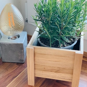 Könnte beinhalten: Ein hölzerner Pflanzkasten mit einem Rosmarinstrauch, neben einer dekorativen Glühbirne. Die Glühbirne hat ein strukturiertes, tropfenförmiges Glas auf einem Betonsockel. Der Pflanzkasten ist aus hellem Holz gefertigt.