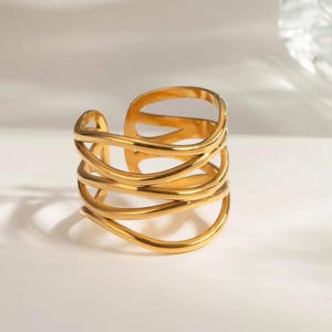 May include: A gold cuff ring with a layered design. The ring has multiple curved bands that wrap around the finger. The open design allows for an adjustable fit. The jewellery is made of a shiny, metallic material.